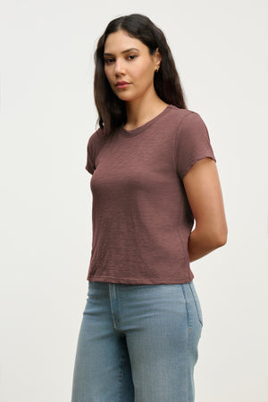 A woman with long dark hair wears the Velvet by Graham & Spencer MARIE TEE in mauve, styled with light blue jeans, standing hands behind her back against a plain background.