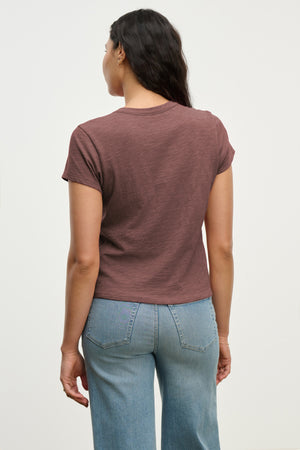 A person with long dark hair, wearing the MARIE TEE by Velvet by Graham & Spencer—a brown cotton slub short-sleeve crewneck t-shirt—and light blue jeans, stands facing away from the camera.