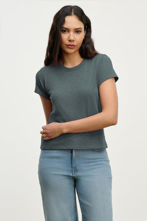 A woman with long dark hair wears the MARIE TEE by Velvet by Graham & Spencer—a short-sleeve dark grey cotton slub t-shirt—paired with light blue jeans, standing against a plain light background. Ideal for any casual occasion.