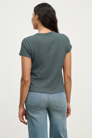 A person stands facing away, wearing the MARIE TEE by Velvet by Graham & Spencer—a cotton slub dark gray t-shirt with a relaxed boxy fit—paired with light blue jeans, against a plain light background.
