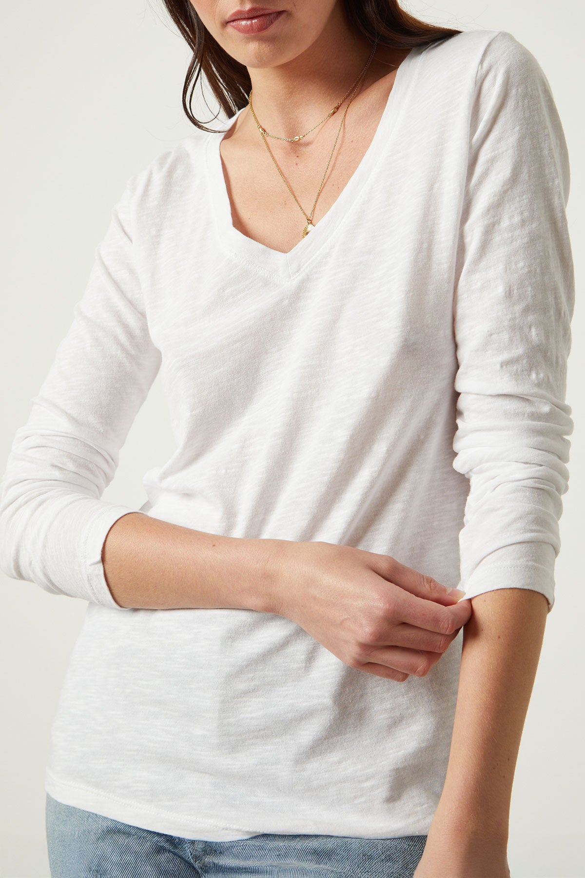 A woman in the BLAIRE TEE by Velvet by Graham & Spencer—a long sleeve white cotton slub V-neck—wears layered gold necklaces and gently touches her opposite sleeve.
