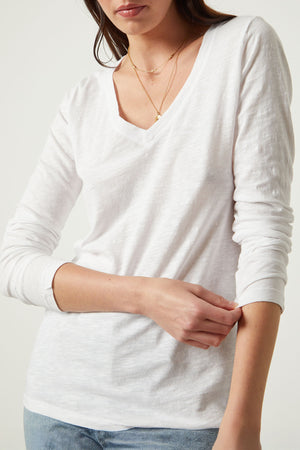 A woman in the BLAIRE TEE by Velvet by Graham & Spencer—a long sleeve white cotton slub V-neck—wears layered gold necklaces and gently touches her opposite sleeve.