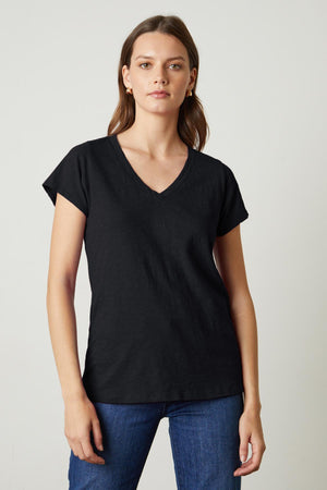 A woman with long hair wearing a black JILIAN TEE from Velvet by Graham & Spencer made from cotton slub fabric and classic blue jeans stands against a plain background.