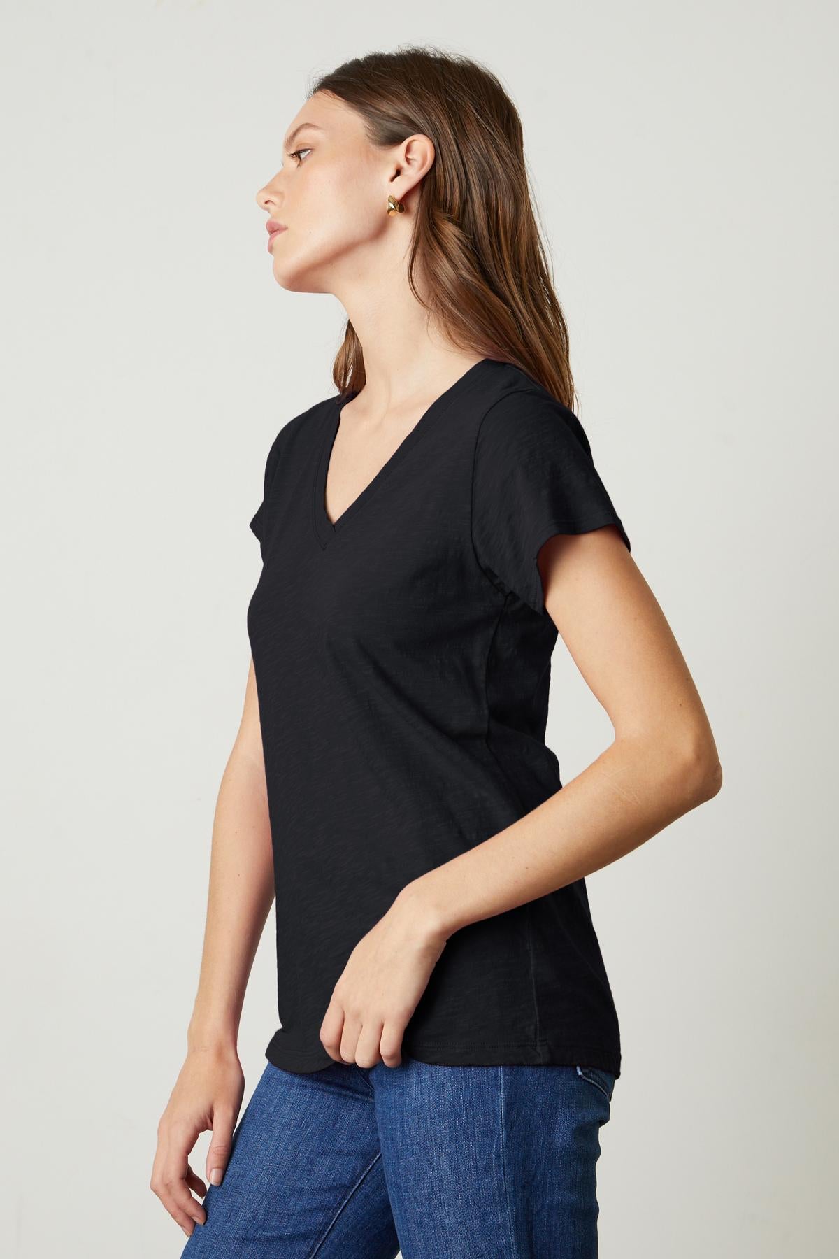 A person with long hair wearing a black JILIAN TEE by Velvet by Graham & Spencer made of cotton slub fabric and blue jeans stands against a plain background, looking to the side.-35629753237697