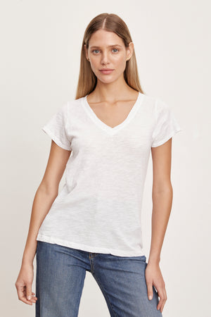 A person with long hair wears a white JILIAN TEE by Velvet by Graham & Spencer made of cotton slub fabric and blue jeans, standing against a plain background.