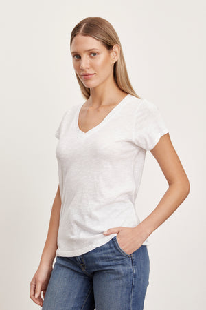 A person with straight blonde hair, wearing a white JILIAN TEE from Velvet by Graham & Spencer made from cotton slub fabric and blue jeans, stands against a plain background.