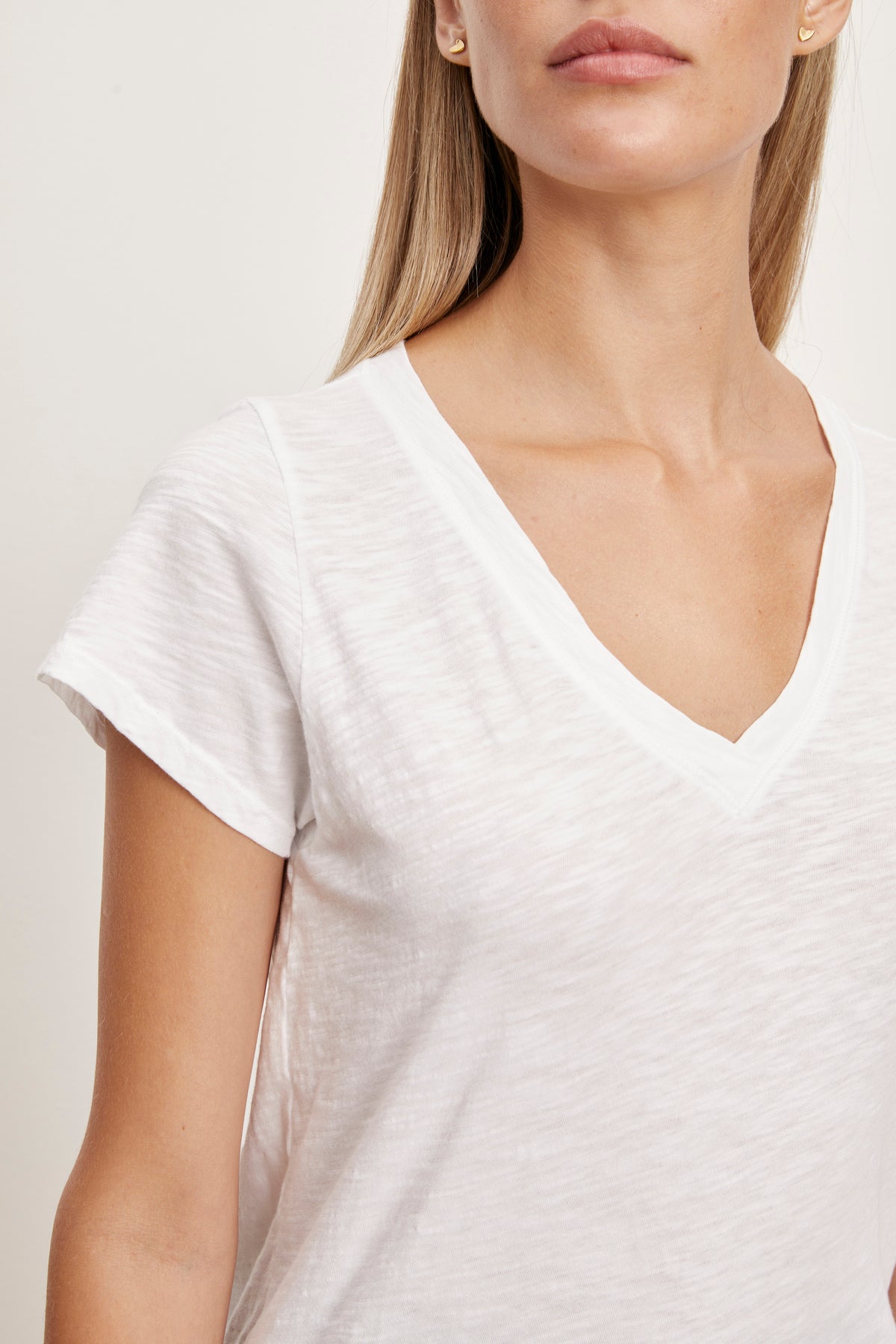 A close-up of a person wearing a white short-sleeve V-neck JILIAN TEE made from cotton slub fabric by Velvet by Graham & Spencer. The person's face is partially visible, looking slightly to the right. They have long blonde hair. This versatile piece is a true wardrobe staple.