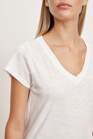 A close-up of a person wearing a white short-sleeve V-neck JILIAN TEE made from cotton slub fabric by Velvet by Graham & Spencer. The person's face is partially visible, looking slightly to the right. They have long blonde hair. This versatile piece is a true wardrobe staple.