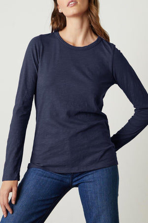 A person wearing a long-sleeved, navy blue LIZZIE TEE by Velvet by Graham & Spencer made of textured cotton slub with a classic crew neckline and jeans stands against a plain light background.