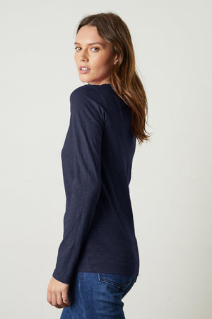 A woman with long brown hair stands sideways against a plain light background, wearing the Velvet by Graham & Spencer LIZZIE TEE in navy textured cotton slub and blue jeans, looking over her shoulder.