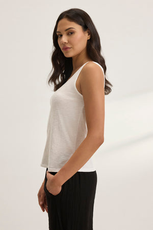 A person with long dark hair stands sideways against a plain background, wearing the AYA TANK TOP from Velvet by Graham & Spencer and black pants, with one hand in their pocket.