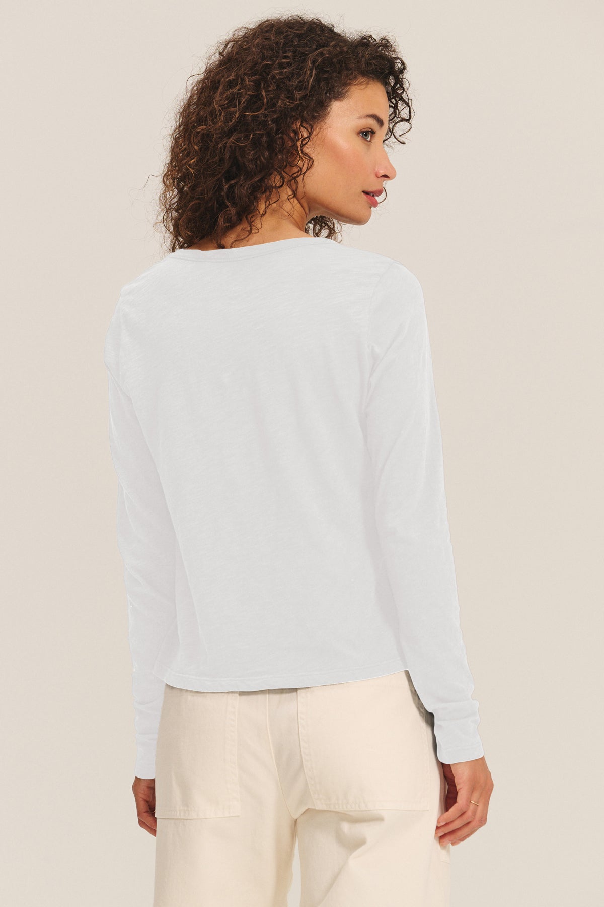 A woman with curly hair faces away, wearing the BECKY TEE by Velvet by Graham & Spencer—a relaxed scoop neck long-sleeve tee in soft cotton slub—paired with light pants against a plain background.-40163382165697