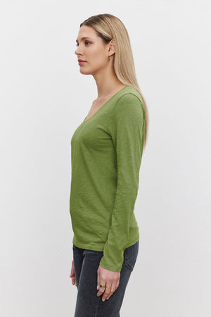 A woman with long blond hair stands in profile, wearing the Velvet by Graham & Spencer BLAIRE TEE in green with dark jeans against a plain white background—styled for effortless everyday wear.