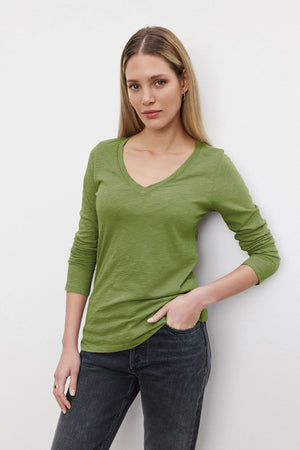 A woman with straight, light brown hair wears the BLAIRE TEE by Velvet by Graham & Spencer—a green, textured cotton slub V-neck long-sleeve shirt—paired with dark jeans, standing against a white background with one hand in her pocket.
