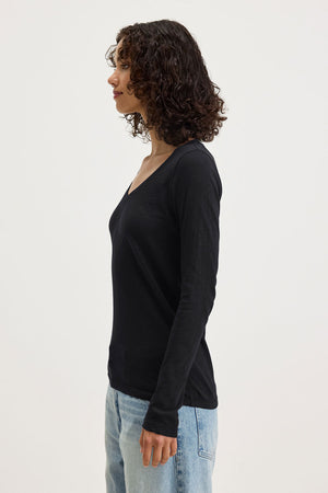 A person with curly hair, in profile against a plain background, wears the BLAIRE TEE by Velvet by Graham & Spencer—a black cotton slub long sleeve V-neck—paired with light blue jeans.