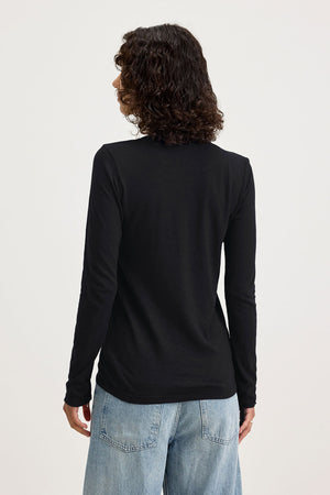 Person stands with their back to the camera, wearing the Velvet by Graham & Spencer BLAIRE TEE— a black long sleeve cotton slub top—paired with light blue jeans, set against a plain light background.