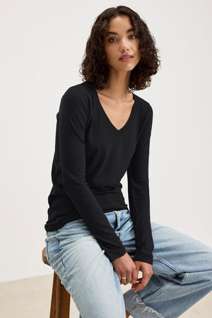 A person with short curly hair wears the Velvet by Graham & Spencer BLAIRE TEE, a long sleeve black V-neck cotton slub top, paired with light blue jeans while sitting on a stool against a plain background.