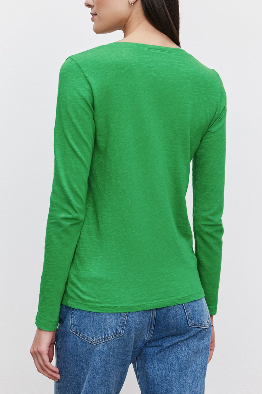 A person with long dark hair is seen from behind, wearing a green BLAIRE TEE by Velvet by Graham & Spencer with long sleeves and blue jeans against a white background.-36201431204033