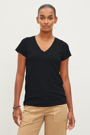 A person stands against a plain background, wearing a black JILIAN TEE crafted from cotton slub fabric by Velvet by Graham & Spencer and tan pants, with one arm resting by their side and the other holding a wrist with a bracelet.
