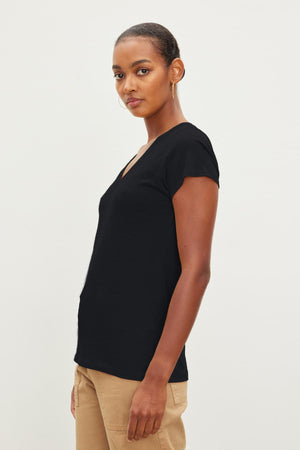 A person with short hair, wearing a black JILIAN TEE by Velvet by Graham & Spencer and beige pants, stands sideways against a plain white background.