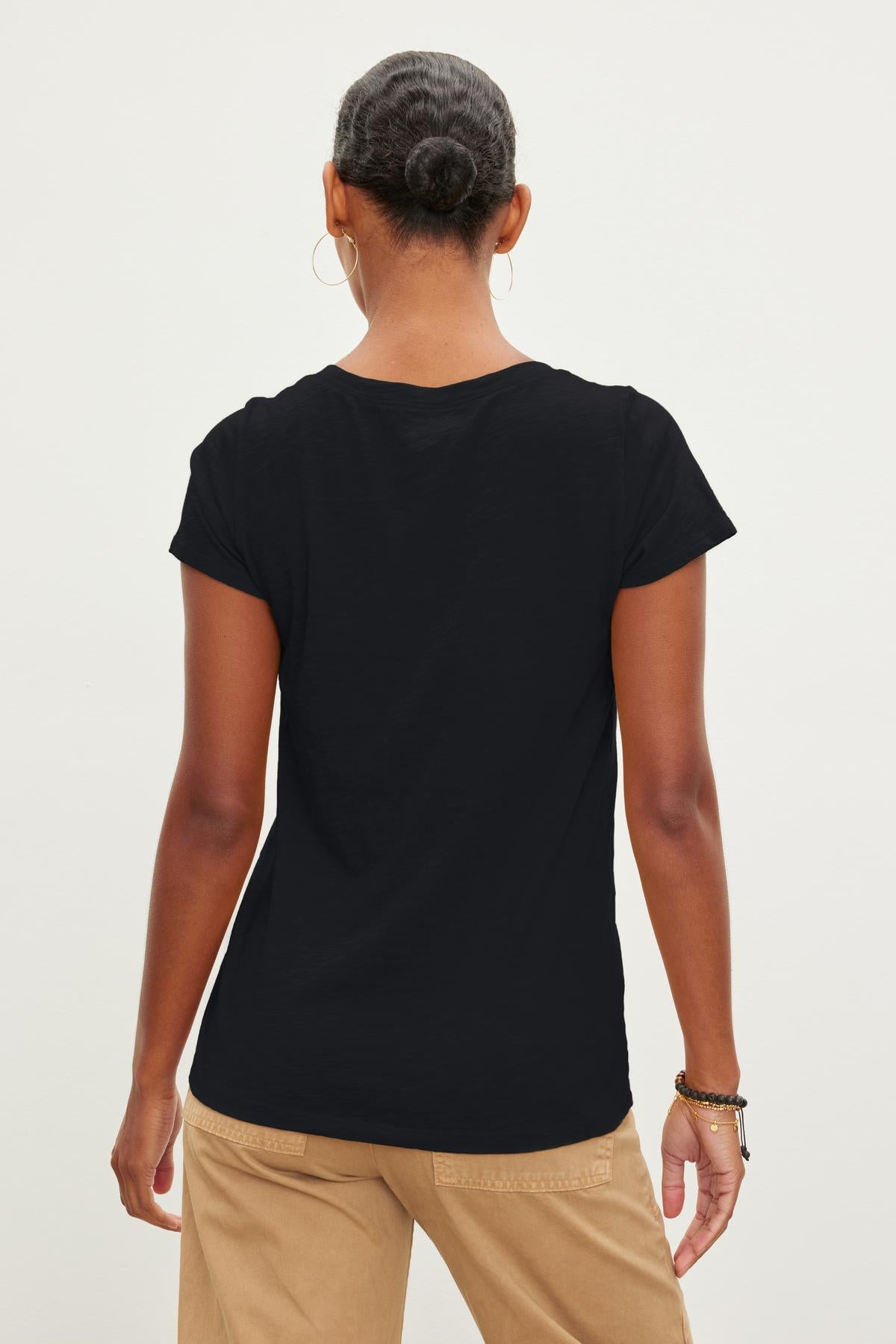 A person with their hair tied back is seen from behind, wearing a plain black JILIAN TEE by Velvet by Graham & Spencer made of cotton slub fabric and beige pants.-35629753204929