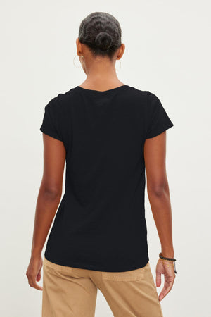 A person with their hair tied back is seen from behind, wearing a plain black JILIAN TEE by Velvet by Graham & Spencer made of cotton slub fabric and beige pants.