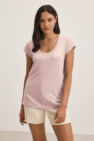 A woman stands against a plain background wearing the Velvet by Graham & Spencer KIRA TEE—a light pink scoop-neck short-sleeve top—paired with cream-colored shorts for an effortless everyday look.