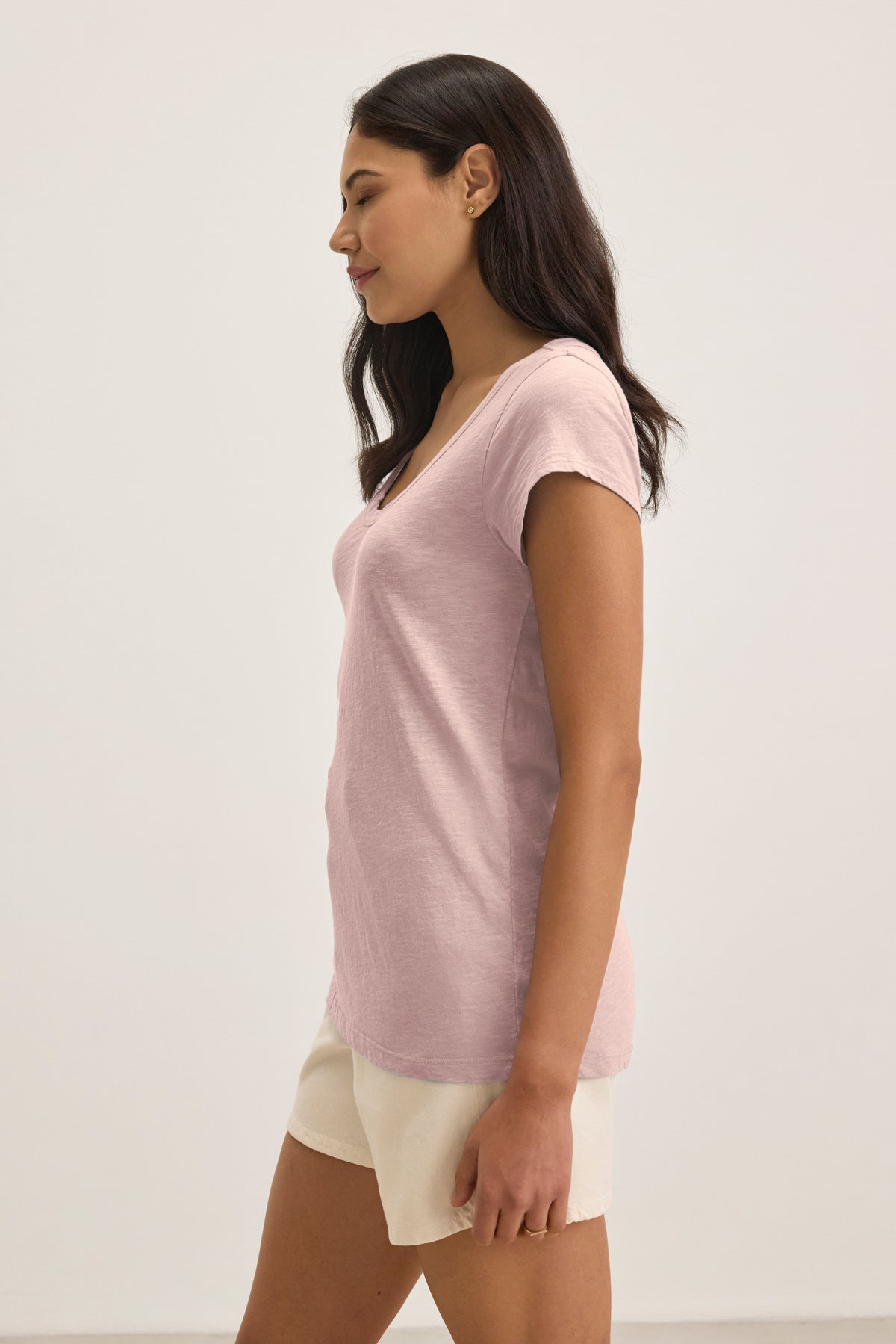 Woman with long dark hair, in profile, wears the KIRA TEE by Velvet by Graham & Spencer—a light pink scoop neck t-shirt—paired with beige shorts against a plain light background.