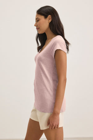 Woman with long dark hair, in profile, wears the KIRA TEE by Velvet by Graham & Spencer—a light pink scoop neck t-shirt—paired with beige shorts against a plain light background.