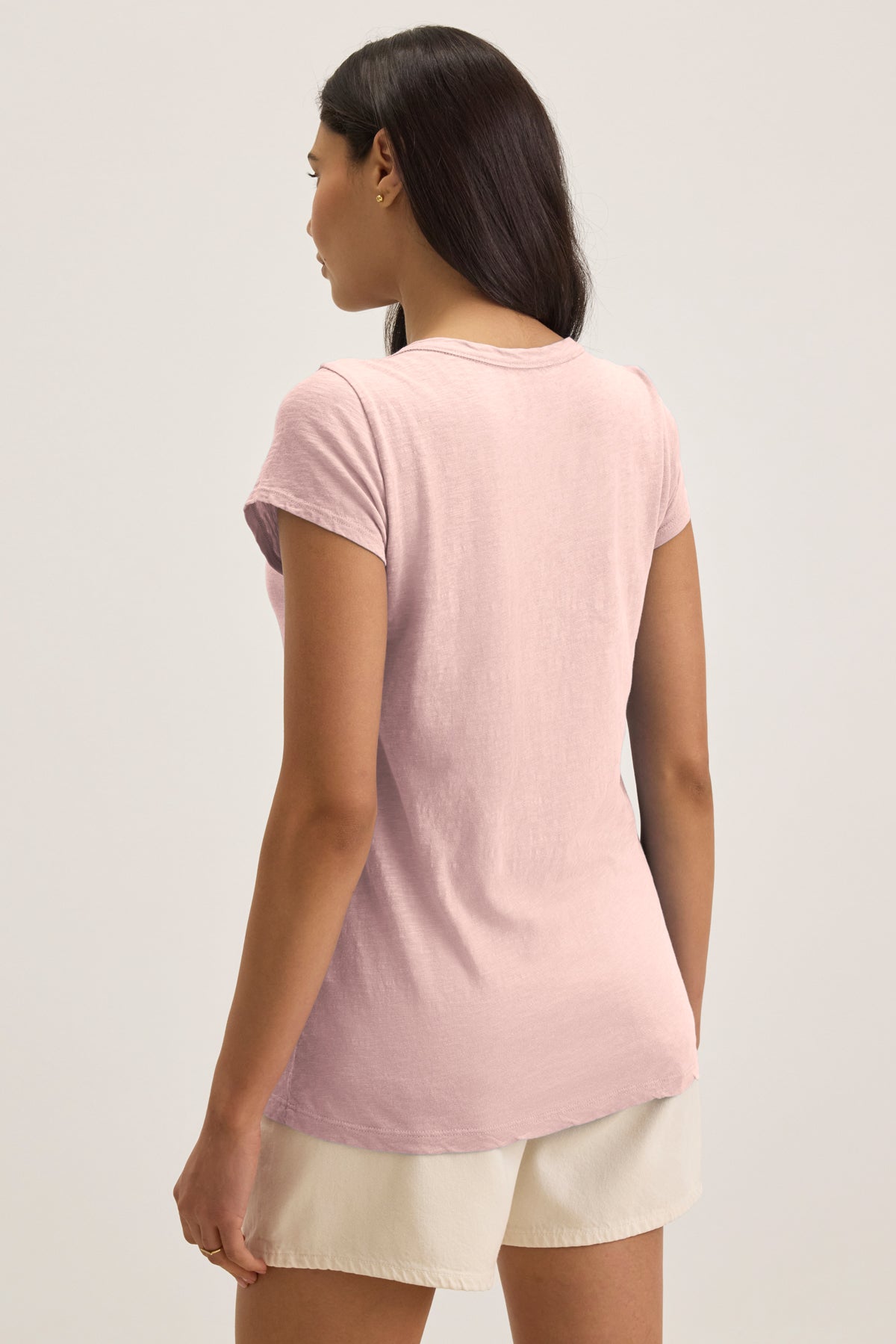 A woman stands facing away, wearing the KIRA TEE by Velvet by Graham & Spencer—a light pink cotton slub scoop-neck t-shirt—paired with light beige shorts against a plain background.