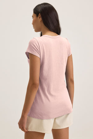 A woman stands facing away, wearing the KIRA TEE by Velvet by Graham & Spencer—a light pink cotton slub scoop-neck t-shirt—paired with light beige shorts against a plain background.