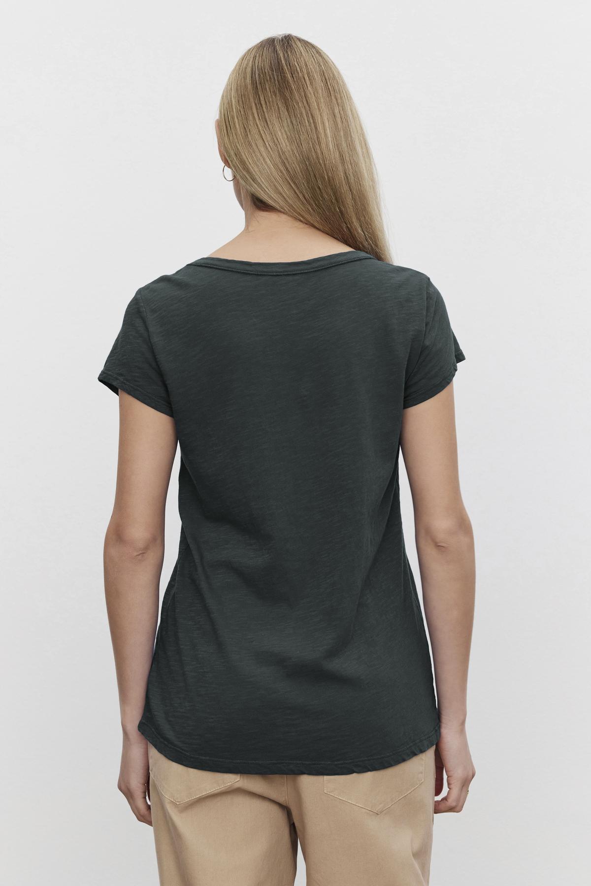 Rear view of a person with long blonde hair wearing the KIRA TEE, a dark green cap sleeve T-shirt by Velvet by Graham & Spencer, paired with beige pants against a white background.-38139482472641