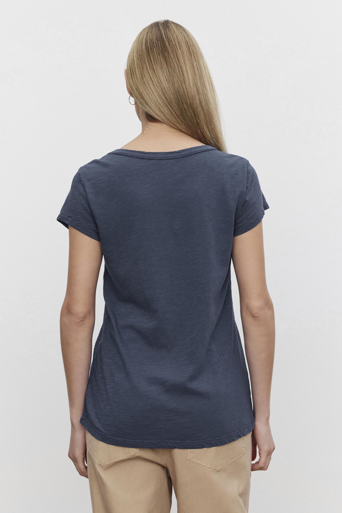 A person with long blonde hair stands facing away, wearing the Velvet by Graham & Spencer KIRA TEE in dark gray with a scoop neckline and beige pants, showcasing their everyday wear.-37606513737921