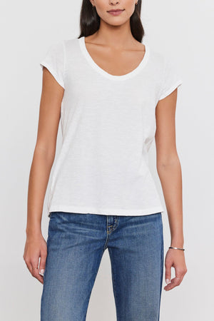 A person with long dark hair, wearing a white short-sleeve V-neck KIRA TEE from Velvet by Graham & Spencer made from a soft cotton slub fabric and blue jeans, stands against a plain white background.