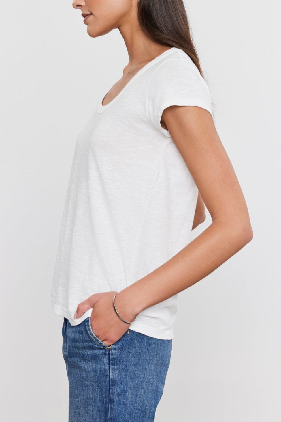 Side profile of a person with long, dark hair wearing a white t-shirt with a scoop neckline and blue jeans, looking forward and placing their left hand in their pocket against a plain white background. Perfected everyday wear crafted from soft cotton slub fabric: the KIRA TEE by Velvet by Graham & Spencer.-35586105966785