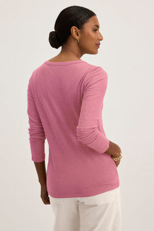A person with dark hair in a bun wears the pink LIZZIE TEE from Velvet by Graham & Spencer, featuring a classic crew neckline and long sleeves, paired with white pants, facing sideways against a plain background.