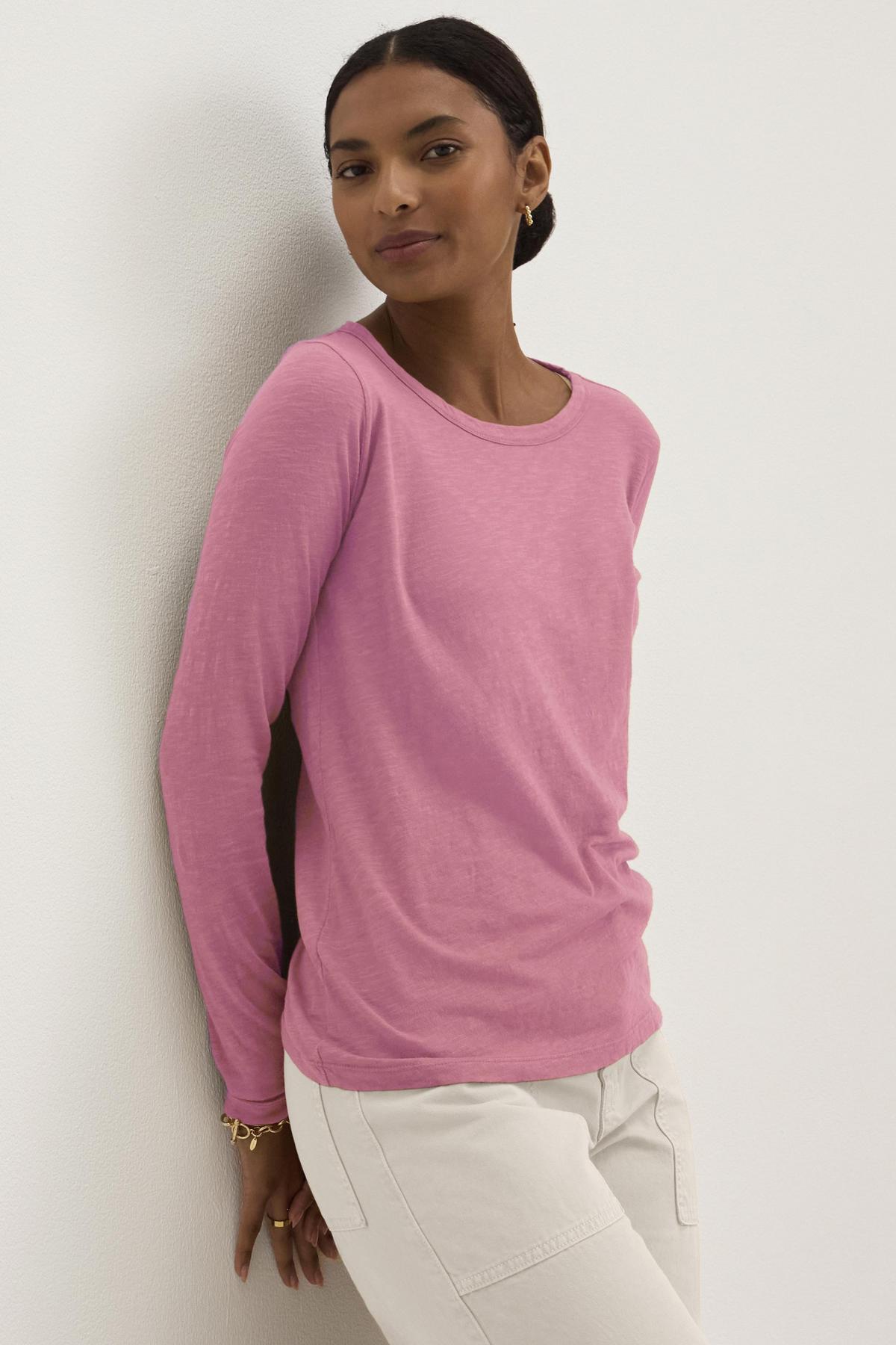 A person wearing the Velvet by Graham & Spencer LIZZIE TEE in pink cotton slub, paired with white pants, leans against a white wall while looking at the camera.-38928750936257