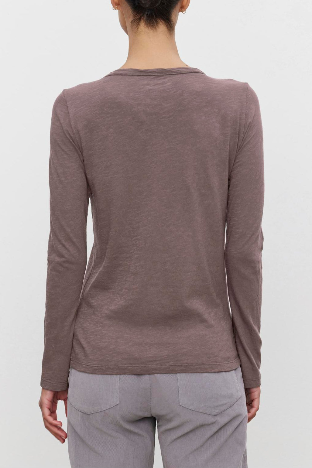 A person with dark hair in a bun is wearing the LIZZIE TEE from Velvet by Graham & Spencer, a long-sleeved brown shirt with a classic crew neckline, paired with light-colored pants, standing with their back to the camera against a plain white background.-37935126085825