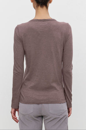 A person with dark hair in a bun is wearing the LIZZIE TEE from Velvet by Graham & Spencer, a long-sleeved brown shirt with a classic crew neckline, paired with light-colored pants, standing with their back to the camera against a plain white background.