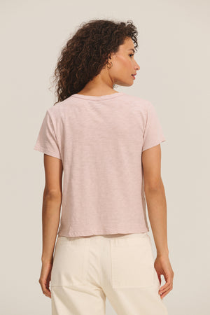 A woman with curly hair is seen from behind wearing the MARIE TEE by Velvet by Graham & Spencer in light pink and off-white relaxed-fit pants, standing against a plain background.