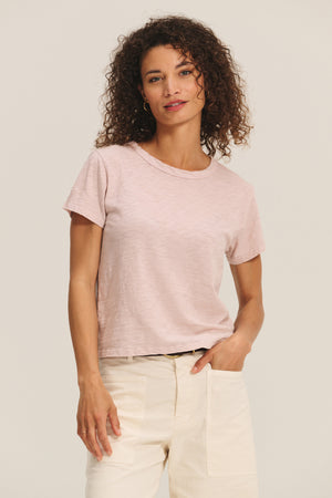 A woman with curly hair wears the Velvet by Graham & Spencer MARIE TEE, a light pink cotton slub short-sleeve shirt in a relaxed boxy fit, paired with cream pants. She stands against a light background, creating an effortless casual look.