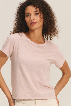 A woman with curly hair wears the Velvet by Graham & Spencer MARIE TEE—a light pink relaxed boxy-fit cotton slub short-sleeve t-shirt—paired with off-white pants, against a plain light background. Perfect for any casual occasion.