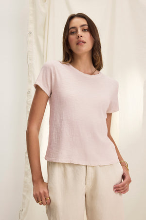 A person wears the MARIE TEE by Velvet by Graham & Spencer in light pink, featuring a relaxed boxy fit and short sleeves, paired with cream-colored pants against a neutral background.