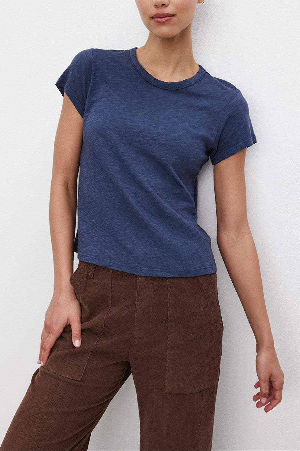 A person with dark hair tied back is wearing the MARIE TEE by Velvet by Graham & Spencer, a navy blue cotton slub T-shirt, paired with brown pants and standing against a plain white background. The outfit's boxy fit makes it perfect for a casual occasion.