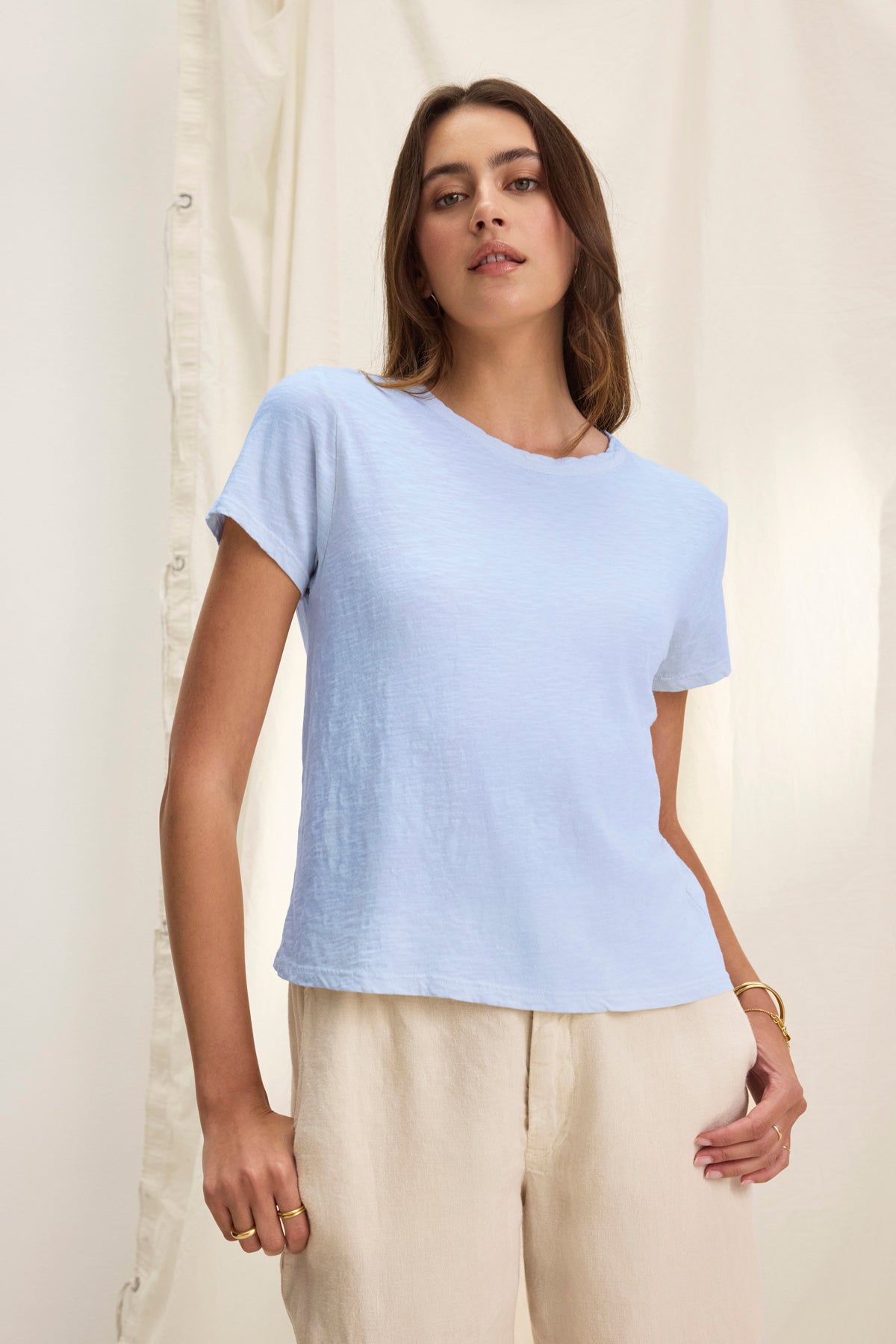 A woman in the MARIE TEE by Velvet by Graham & Spencer—a light blue cotton slub short-sleeve t-shirt with a relaxed boxy fit—pairs it with beige pants, standing before a cream backdrop, ideal for casual occasions.-39319770267841