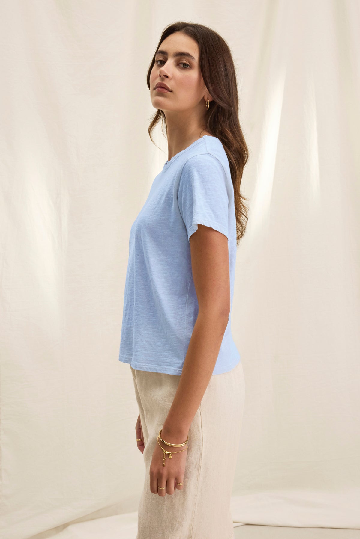 A woman stands sideways, wearing the Velvet by Graham & Spencer MARIE TEE—a light blue cotton slub short-sleeve t-shirt with a relaxed boxy fit—and beige pants, minimal jewelry, against a neutral fabric backdrop; ideal for casual wear.-39319770300609
