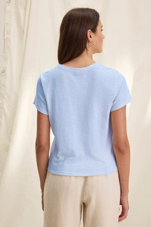 A woman with long brown hair stands with her back to the camera, wearing the MARIE TEE by Velvet by Graham & Spencer in light blue and beige pants against a neutral backdrop—an ideal choice for casual occasions.