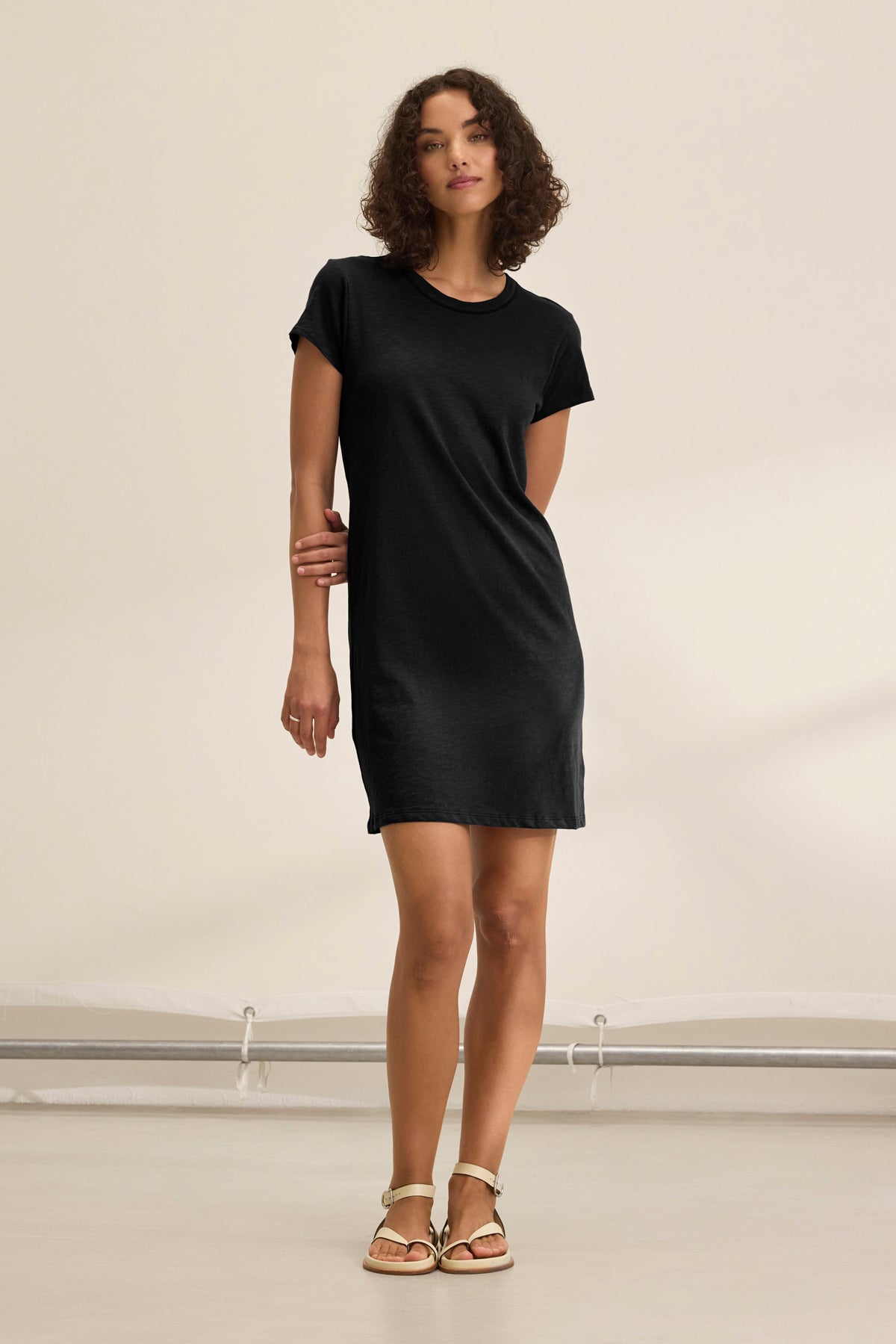 A person stands indoors against a plain, light background, wearing the SIA T-SHIRT DRESS by Velvet by Graham & Spencer—a mini-length black cotton slub dress—paired with beige sandals.