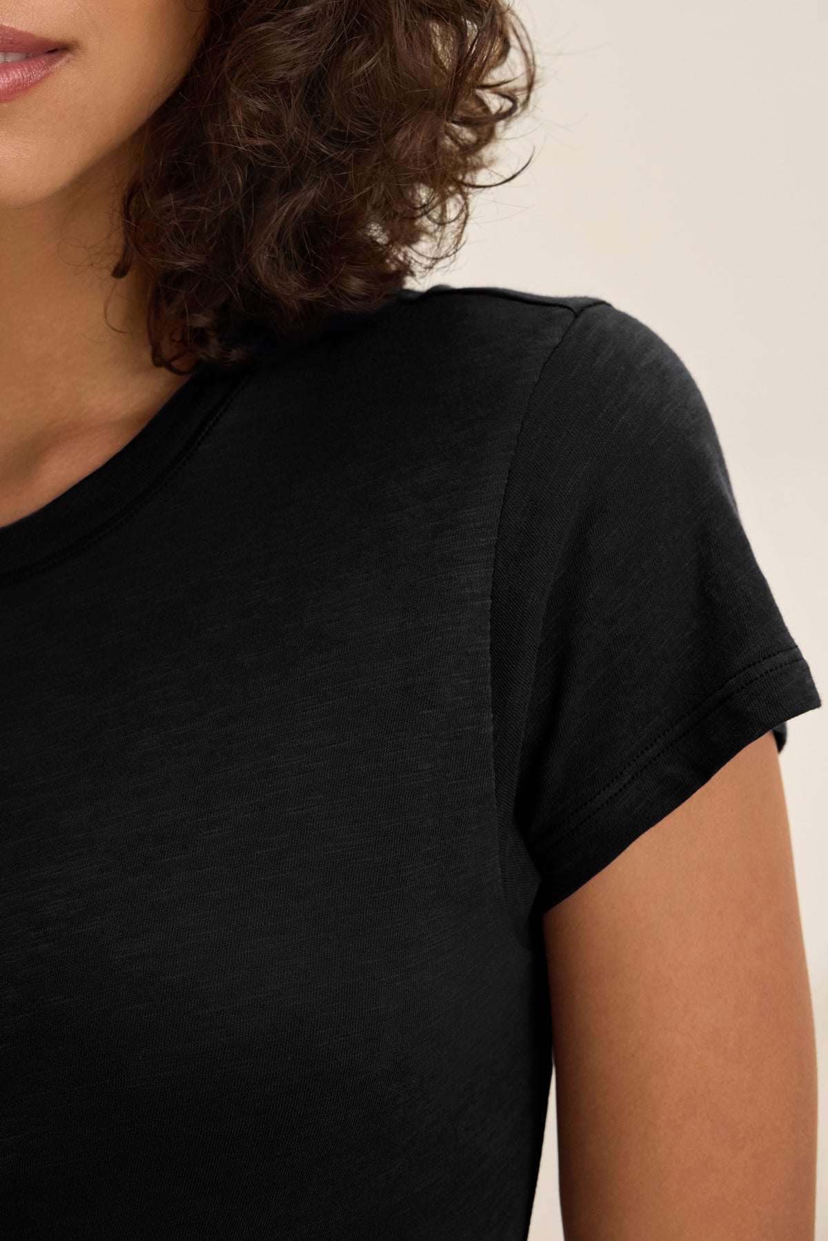 A close-up of a person wearing the Velvet by Graham & Spencer SIA T-SHIRT DRESS, with part of their face, shoulder, and curly brown hair visible against a plain background.-39079428849857
