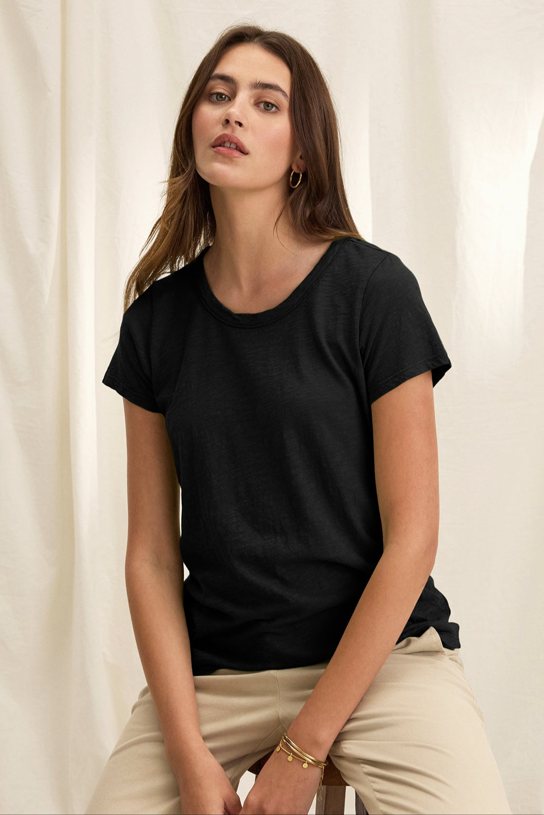 A person sits against a light fabric backdrop, wearing a TILLY TEE by Velvet by Graham & Spencer and beige pants, exuding the relaxed vibe of a California classic with their neutral expression.-38866044846273